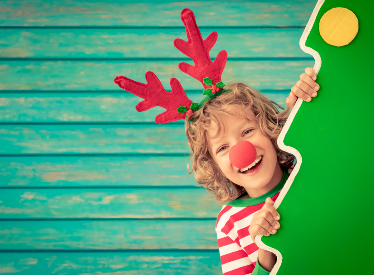 A child with a red nose and antlers, peeking out from behind a Christmas tree.