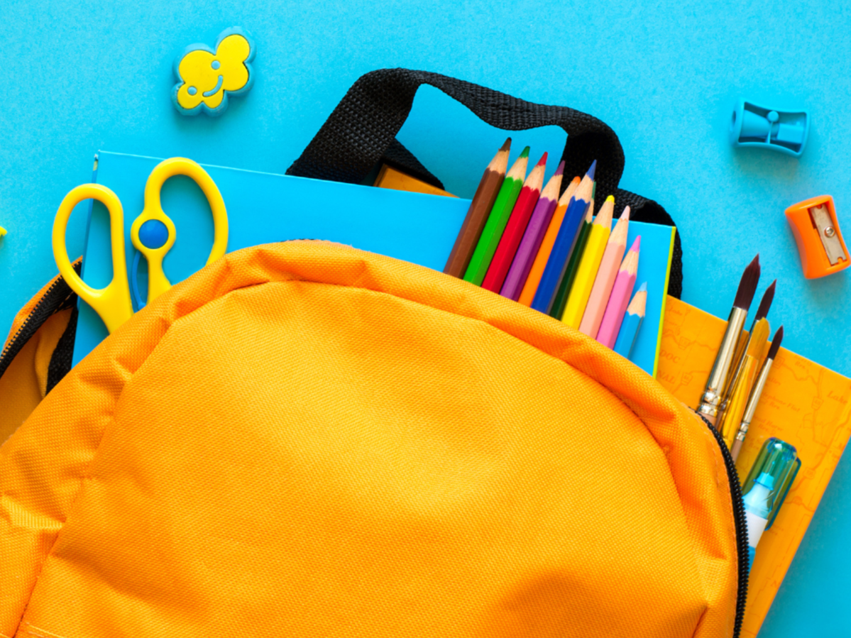A yellow backpack with school supplies pouring out of it on a light blue background.