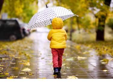 Child in a yellow jacket with a white and black polka dot umbrella walking in the rain.