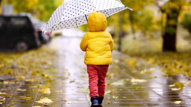 Child in a yellow jacket with a white and black polka dot umbrella walking in the rain.