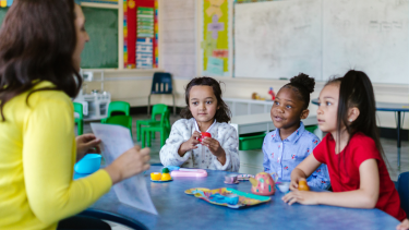 Parent/Guardian Teacher Conferences Children sitting at a rainbow table with a teacher.