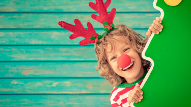 A child with a red nose and antlers, peeking out from behind a Christmas tree.