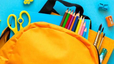 A yellow backpack with school supplies pouring out of it on a light blue background.