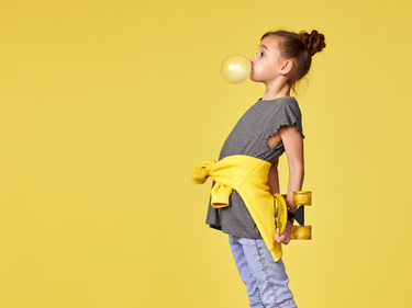 A girl dressed in yellow, blowing a bubble with bubble gum.